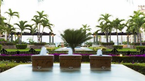A fountain with three stone basins in a landscaped garden with palm trees and a view of a building in the background.