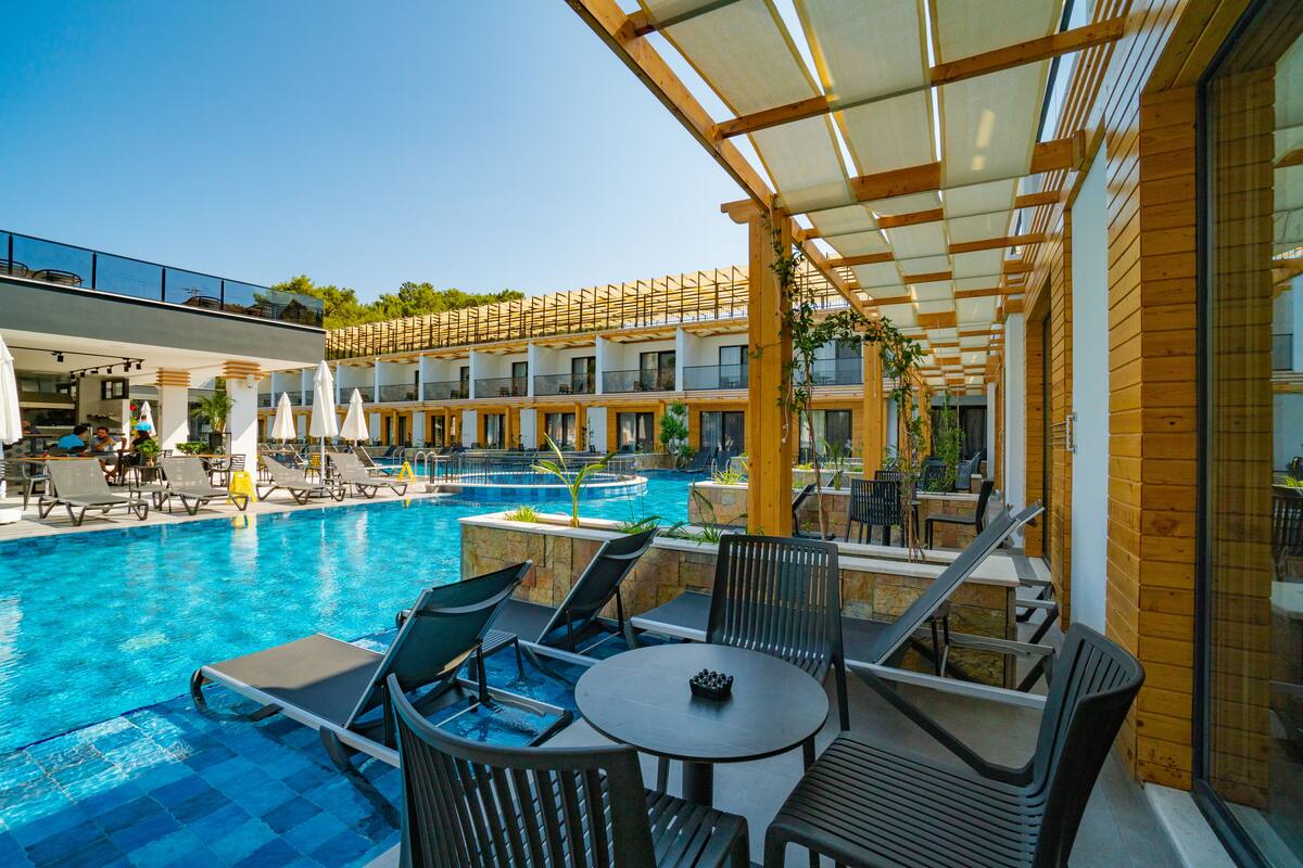 A pool area with lounge chairs and tables, a clear blue pool, and a covered seating area with wooden beams and glass walls.
