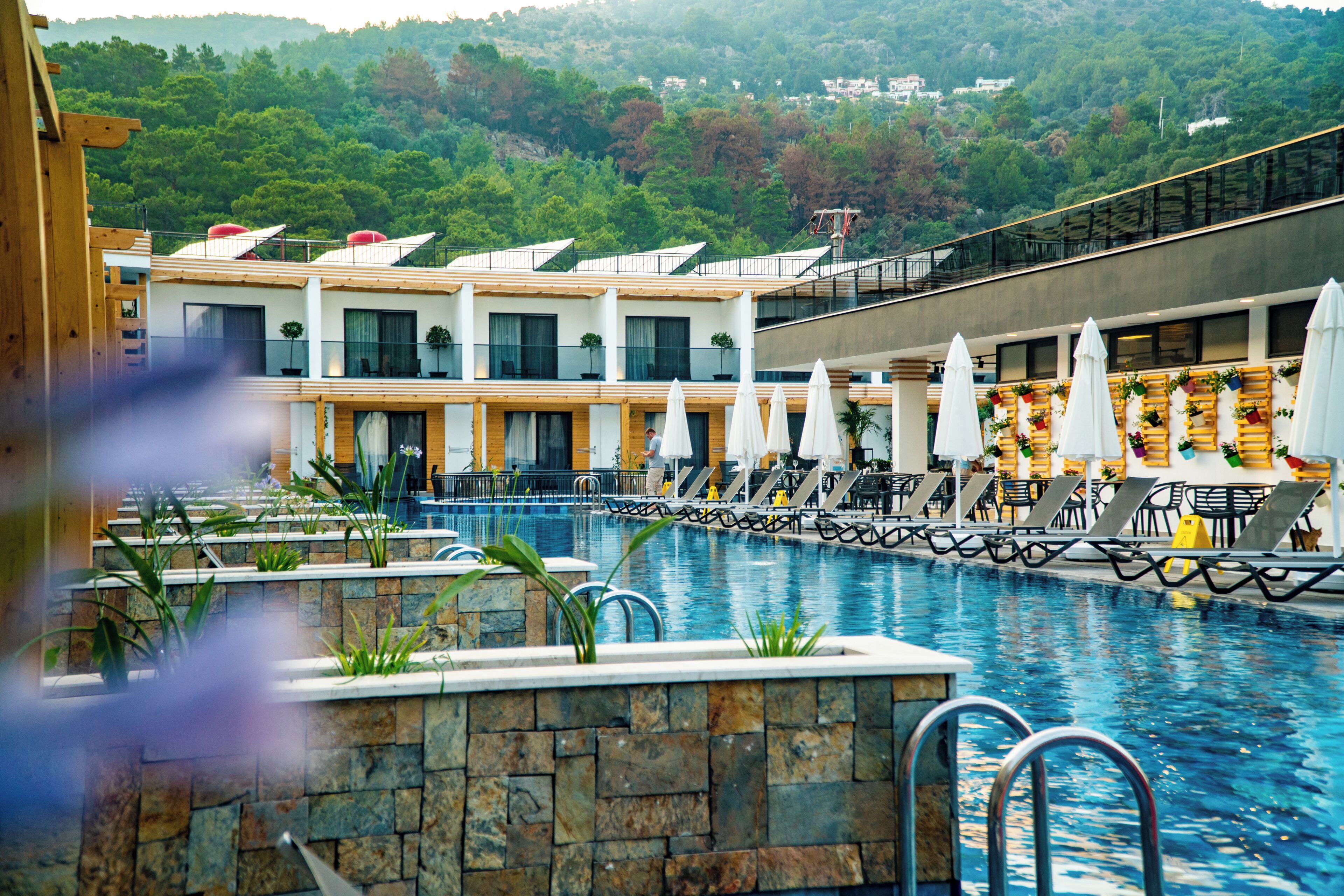 A swimming pool with lounge chairs and umbrellas, surrounded by a stone wall and a building with multiple windows and balconies.