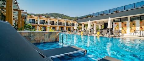 A swimming pool with lounge chairs and umbrellas, surrounded by buildings with balconies and windows overlooking a hilly landscape.