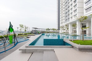 Uma piscina ao ar livre moderna com um deck revestido de azulejos, rodeada por vegetação e um edifício alto ao fundo.