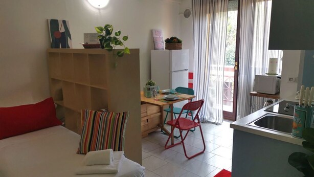 Een compacte kamer met een bed, een bureau, een stoel, een kleine keukenhoek en een glazen schuifdeur naar een buitenruimte.