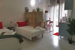 A hotel room with two beds, a desk, a chair, a small table, a shelf with plants, and a window with curtains.