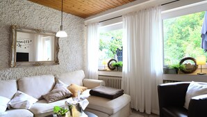 Una sala de estar acogedora con un sofá blanco, un sillón negro y una mesa de centro con una bandeja de bebidas y una planta. Un espejo con un marco decorativo cuelga de la pared, y una ventana con cortinas blancas ofrece vistas al verdor exterior.