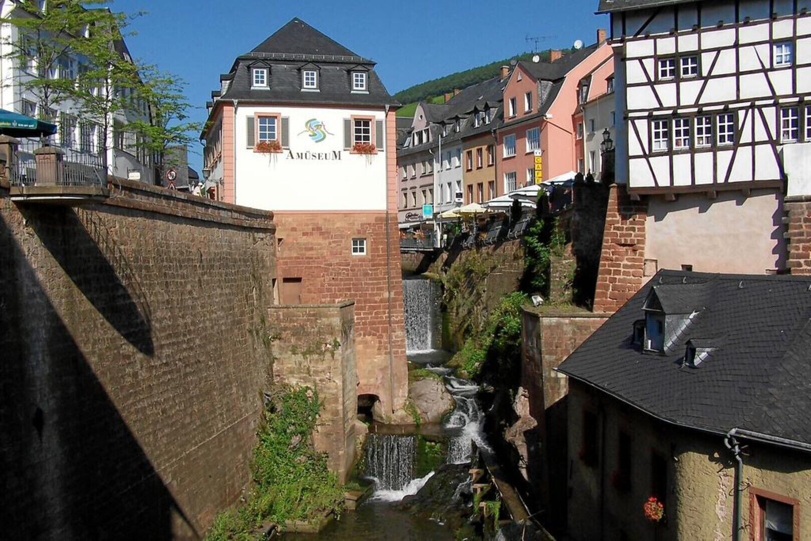 Um edifício com a placa 'AMUSEUM', arquitetura europeia tradicional, uma pequena cascata e uma passagem estreita entre edifícios.