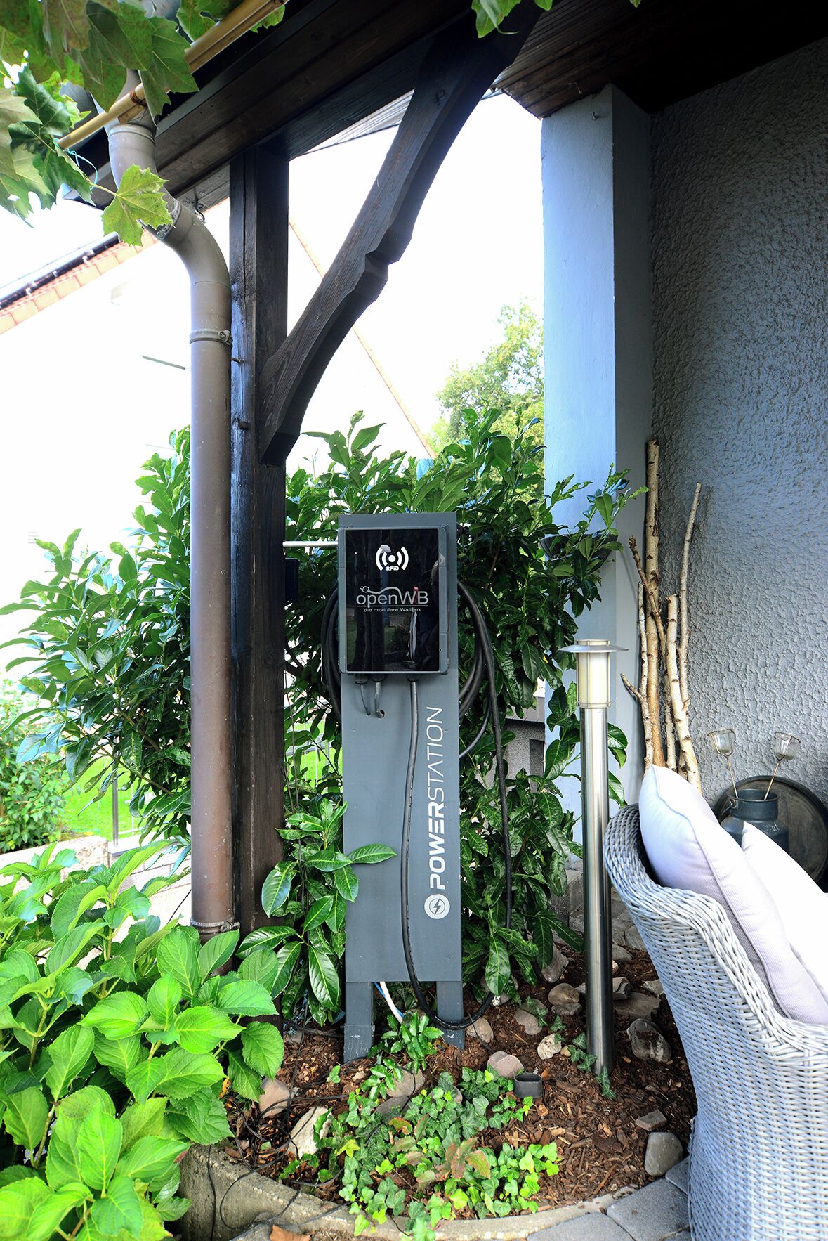 An outdoor electric charging station labeled "openWB" with a power outlet, surrounded by greenery and a wicker chair.