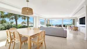 A modern living room with a large dining table, comfortable seating, and a view of the ocean. - Unit 24 - 3 Bed Ocean View (TERRIGAL)