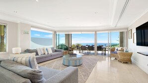 A spacious living room with a large window offering a view of the ocean, a grey sofa, a round coffee table, and a wicker side table. - Unit 24 - 3 Bed Ocean View (TERRIGAL)