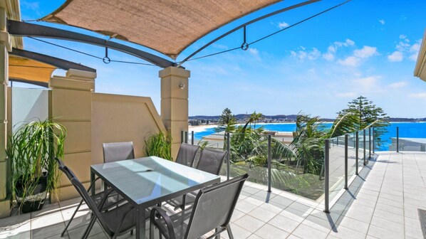 Une terrasse avec une table et des chaises en verre, donnant sur une plage et l’océan.