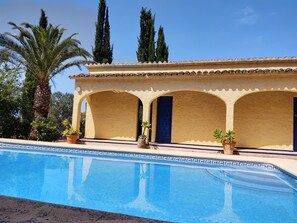 A poolside area with a clear blue pool, a building with a tiled roof, and palm trees in the background.