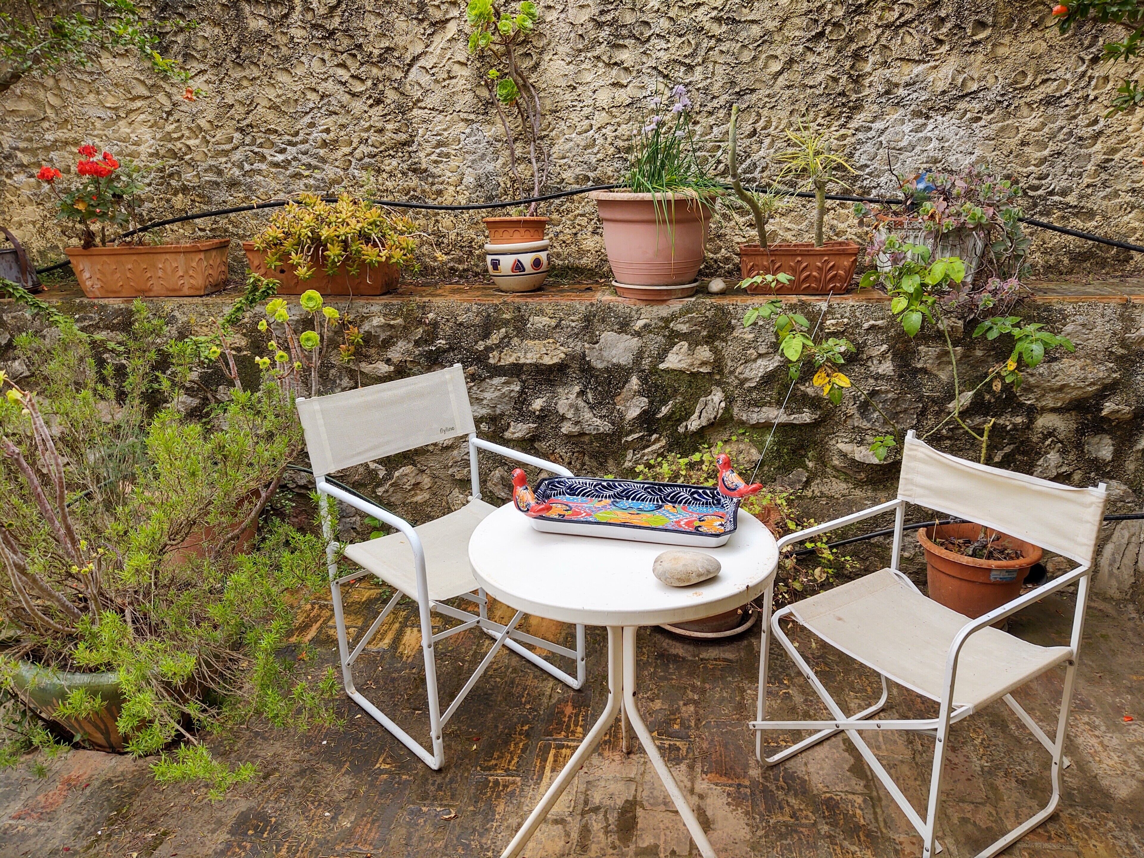 A small outdoor seating area with two white metal chairs and a round table, surrounded by various potted plants and a stone wall.