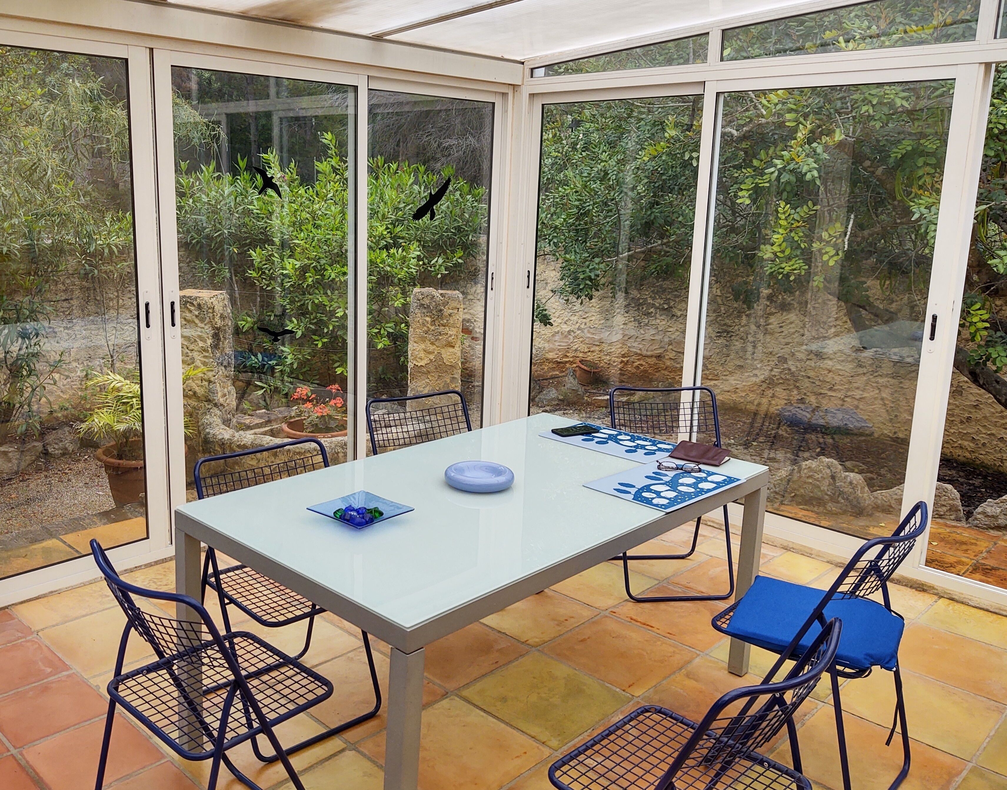 Une véranda avec une table et des chaises en verre, entourée de plantes et d’un mur en pierre.