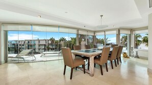 A spacious dining area with a large table and chairs, large windows offering a view of a residential complex and palm trees. - Unit 36 - 4 Bed Platinum Ocean View (TERRIGAL)