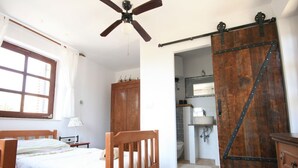 Una camera da letto con un letto di legno, una ventola a soffitto, pavimento in mattoni, una porta scorrevole e un bagno visibile attraverso una porta aperta.