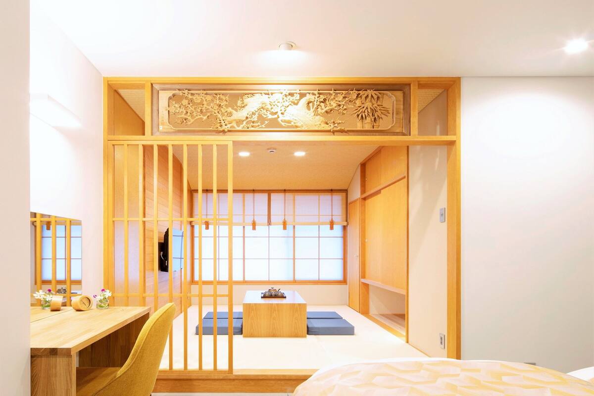 Une pièce de style japonais traditionnel, avec un sol recouvert de tatamis, un petit bureau en bois, un lit avec une couvre-lit à motifs et une porte coulissante ornée d’un panneau décoratif.