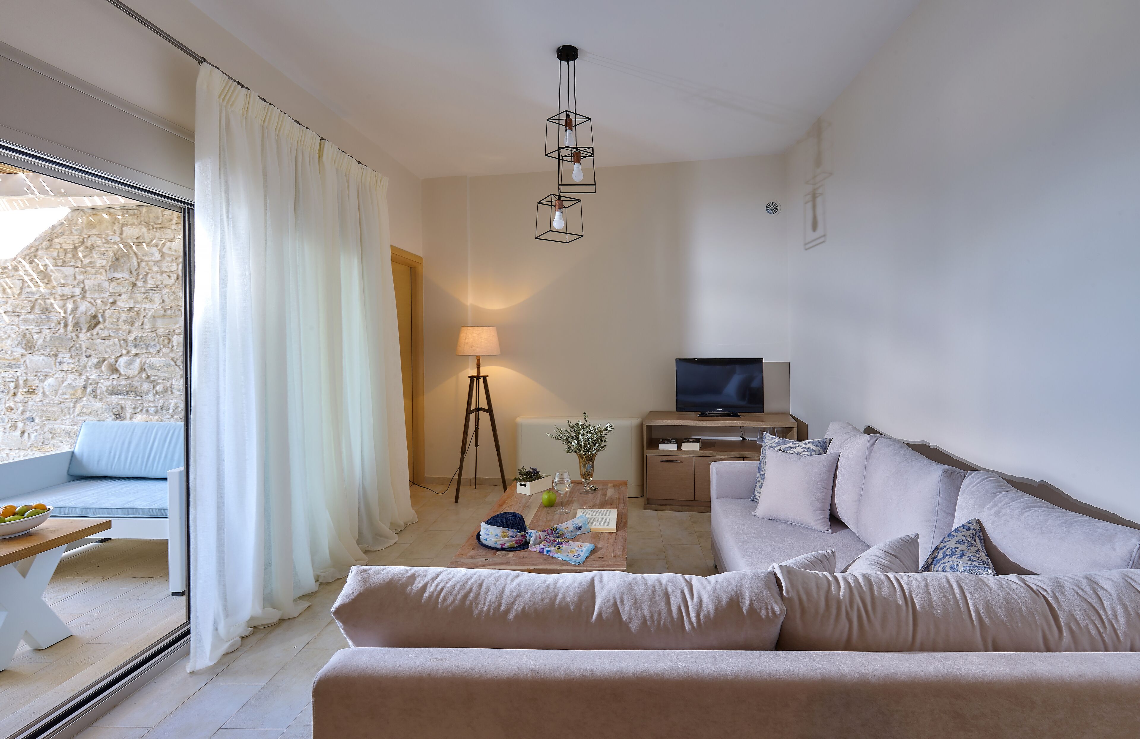 Een moderne woonkamer met een bank, salontafel en televisie. Een stenen muur is zichtbaar door een glazen deur, en er staat een lamp op een driepoot.