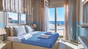 A bedroom with a large bed, a nightstand, a lamp, a window with curtains, and a view of the sea. - Elkysti Crete (Faistos)