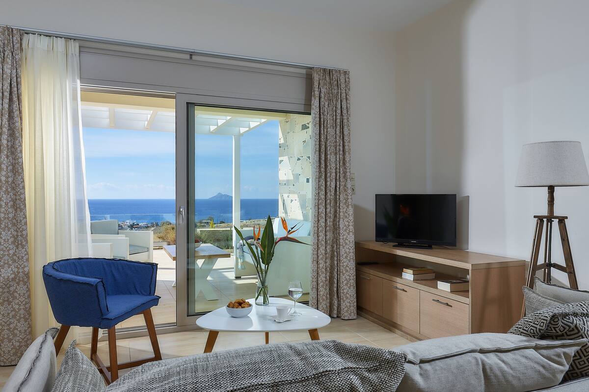 Ein modernes Wohnzimmer mit Fernseher, einer Couch, einem Couchtisch und einem Sessel. Ein Balkon bietet Blick auf das Meer.