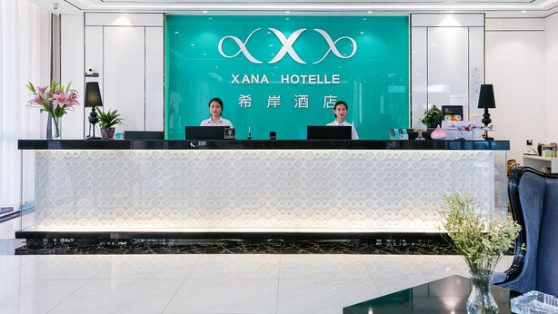 A hotel reception desk with a teal backdrop and a logo, two receptionists, and a seating area with a vase of flowers. - Xi'an Hotel Xiaoshan Airport Branch (Hangzhou)