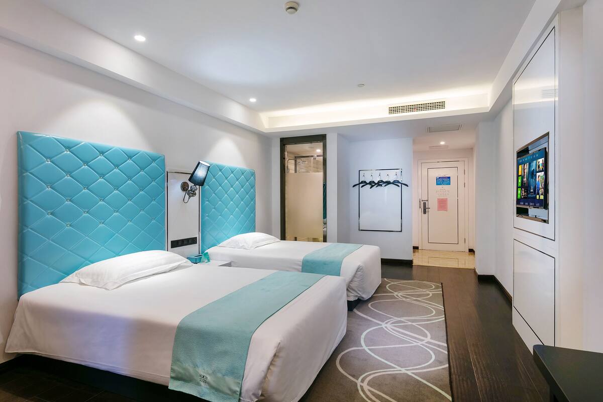 A hotel room with two beds, a large blue headboard, a bedside table with a lamp, a wall-mounted TV, and a door leading to a bathroom.