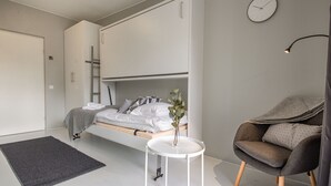 Une chambre à coucher moderne avec un lit, une petite table de chevet blanche, un fauteuil gris et une horloge murale.
