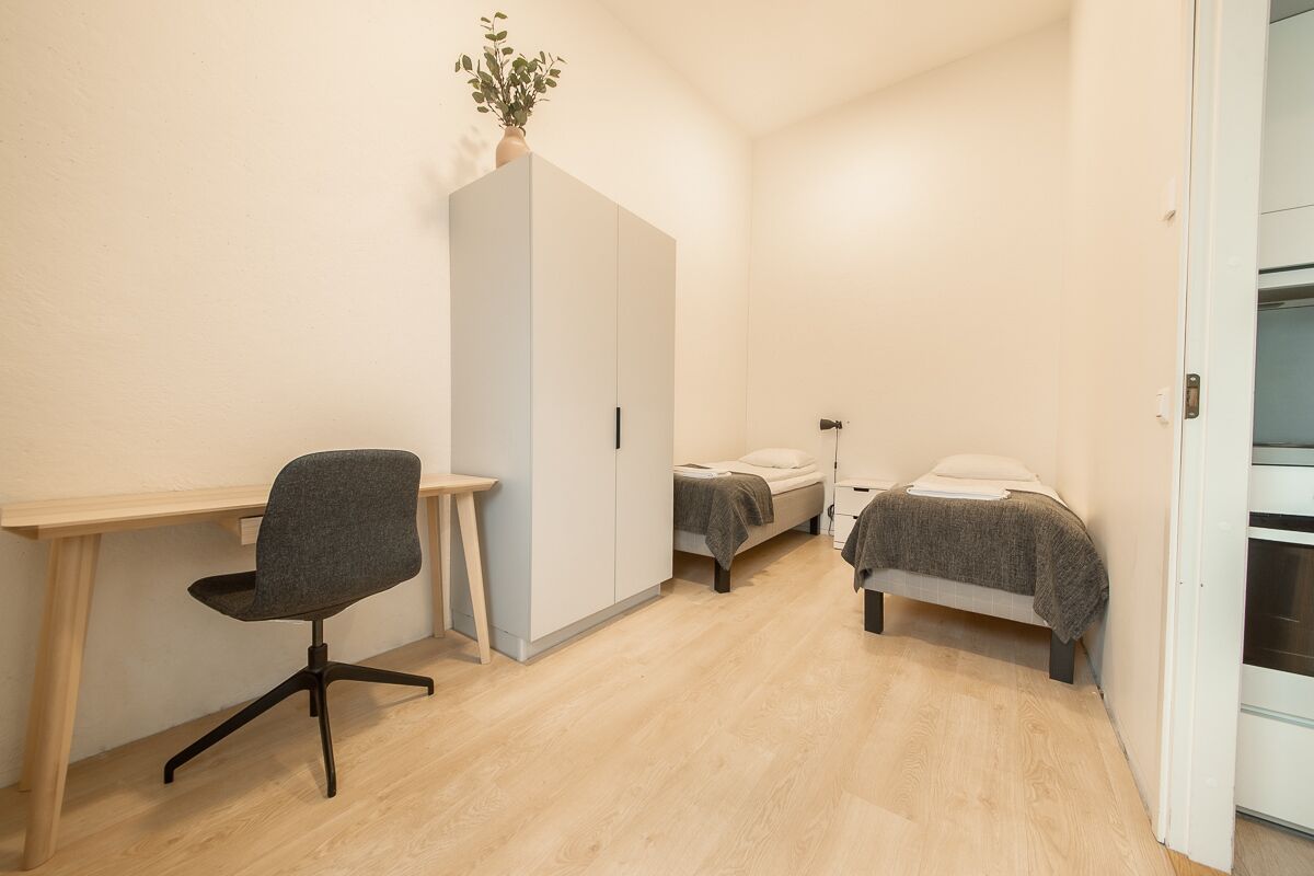 A compact room with a desk, two single beds, a wardrobe, and a small table.
