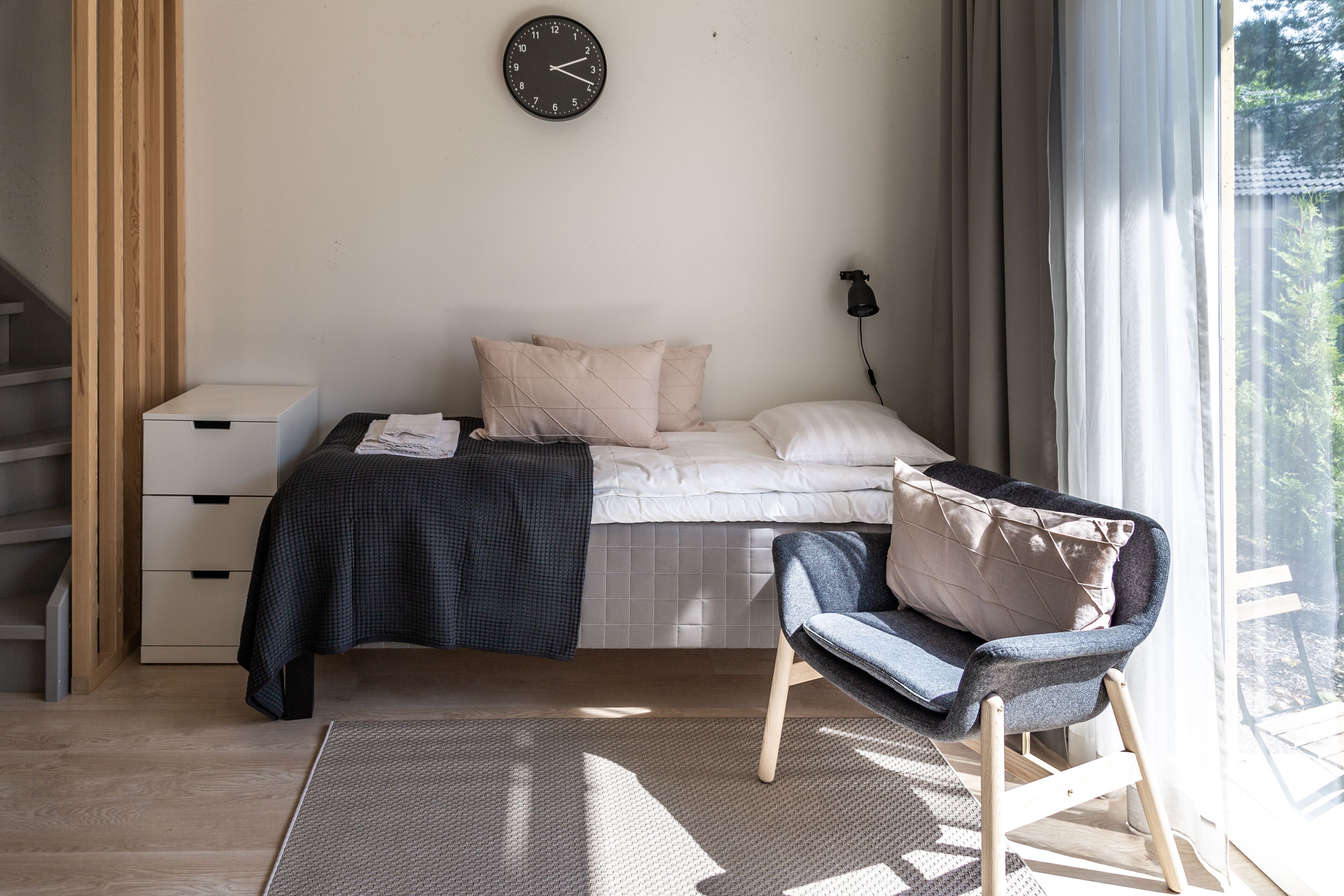 A bedroom with a bed, a chair, a bedside table, a clock, and a window with curtains.