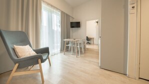 Un soggiorno moderno con una poltrona grigia, un tavolo da pranzo con sedie, un televisore a schermo piatto montato a parete e una cucina visibile attraverso una porta aperta.