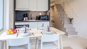 A modern kitchen with white cabinets, a black microwave, and a coffee maker. There is a dining table with white chairs and a fruit bowl. A staircase with a glass railing leads to an upper level. - Spot Apartments Airport (Vantaa)