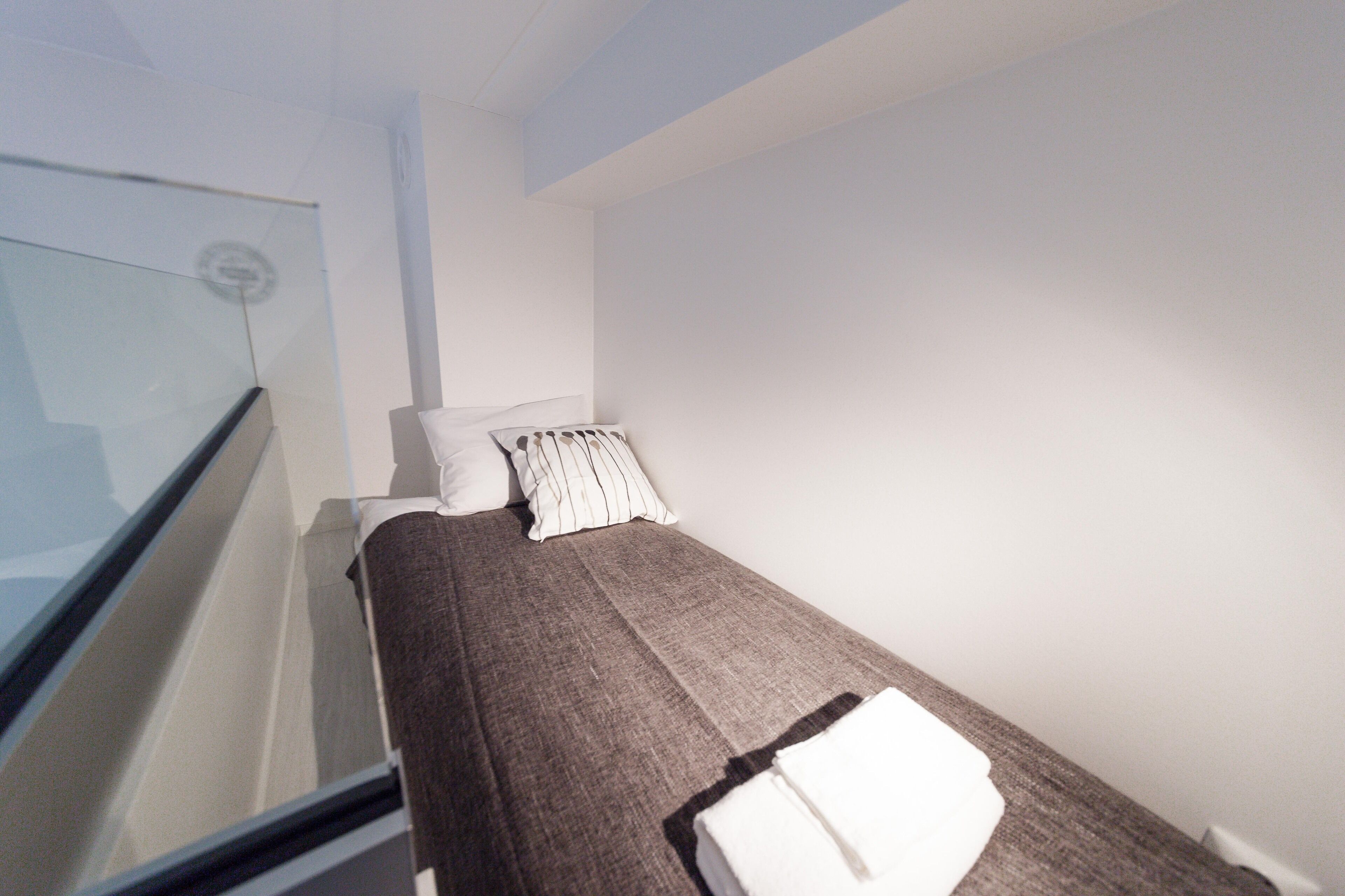 Een enkel bed met een bruine dekbedovertrek en witte kussens in een kamer met witte wanden en een glazen deur.