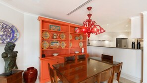 A dining area with a wooden table and chairs, a red cabinet with decorative plates, a red chandelier, and a kitchen in the background. - Unit 33 - 3 Bed Garden View (TERRIGAL)