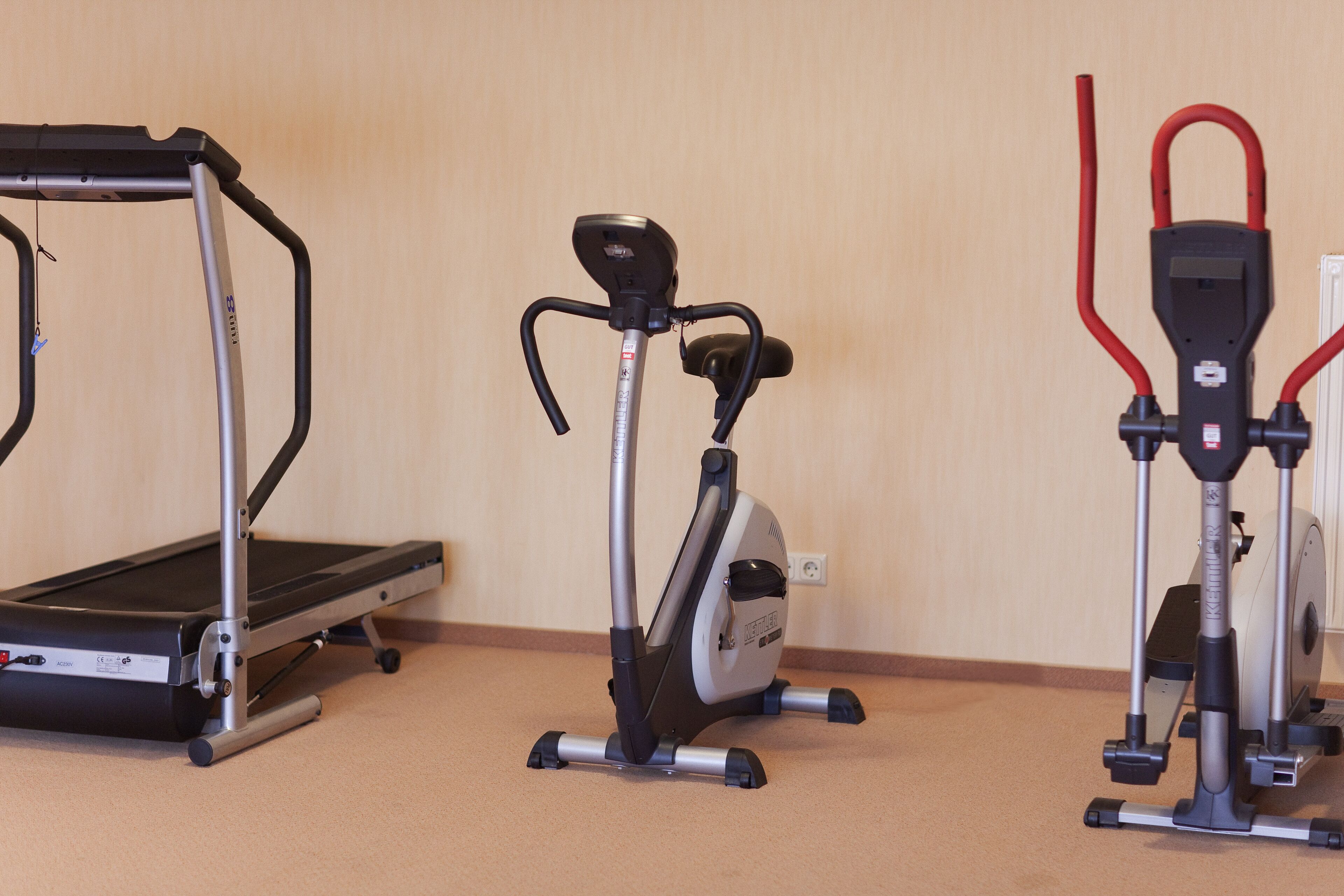 Three stationary exercise machines in a room with a carpeted floor and a plain wall.