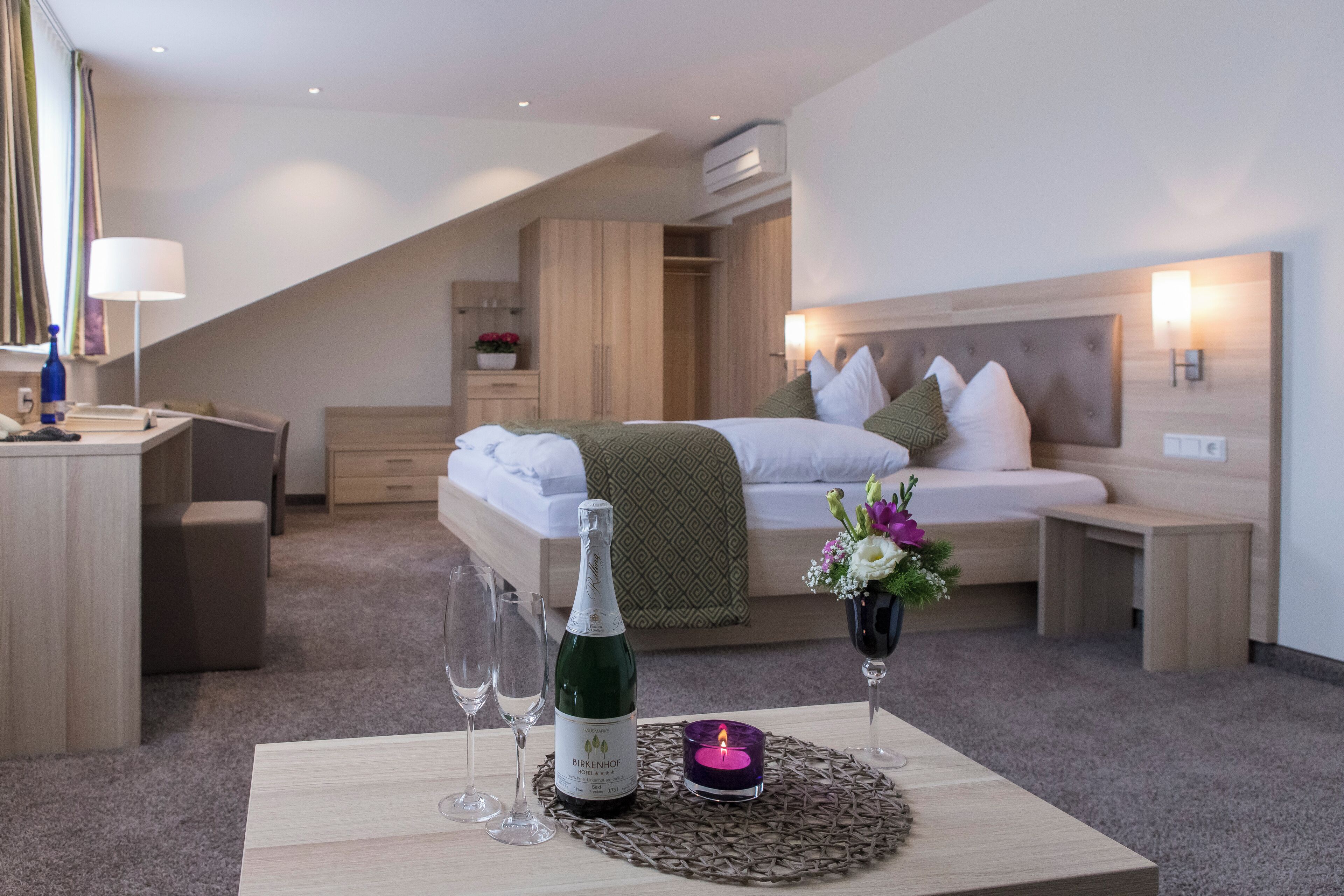 A modern hotel room with a bed, bedside table, and a desk with a lamp.