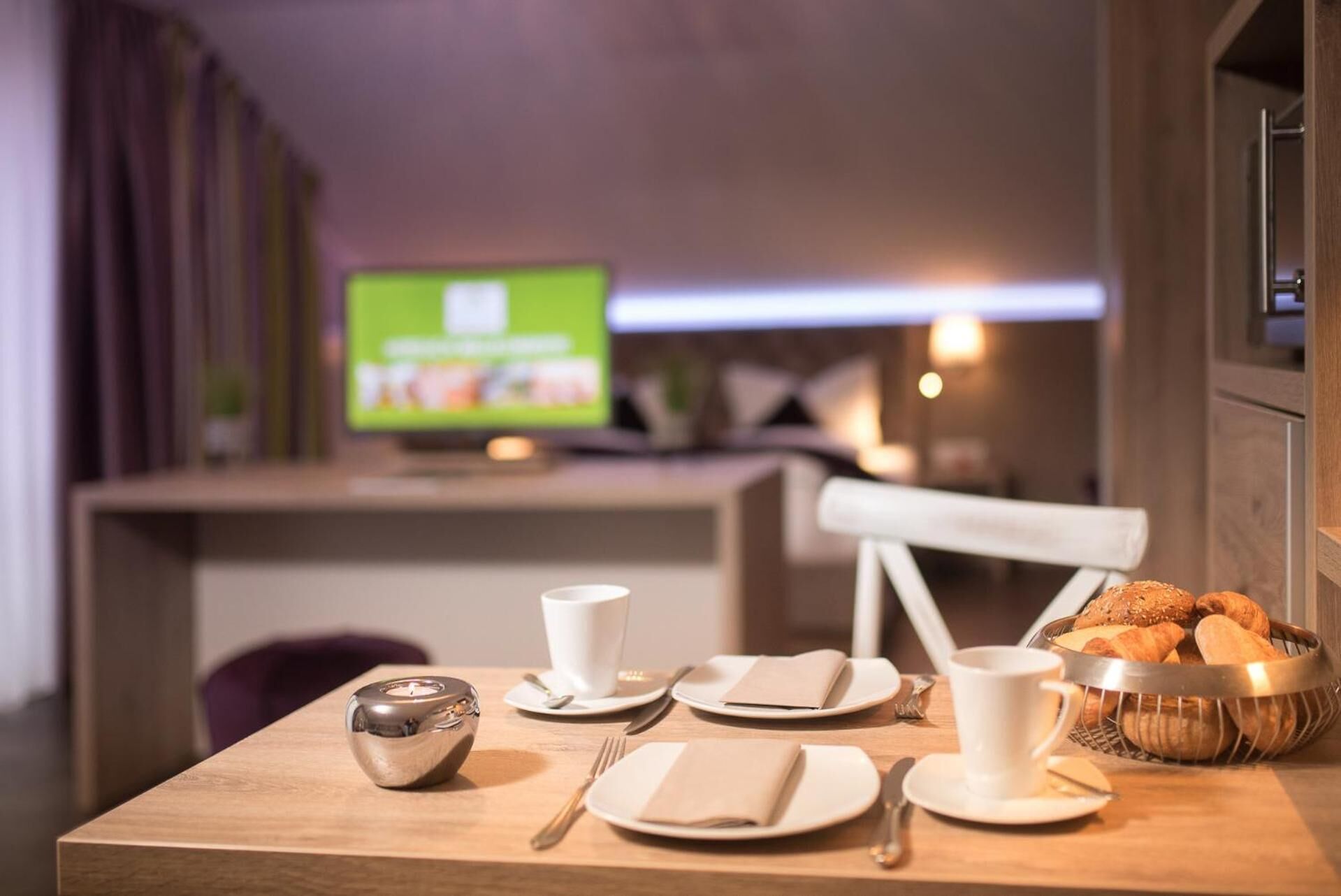 A neatly set dining table with plates, cups, and a bread basket, a small metallic object, and a television in the background.