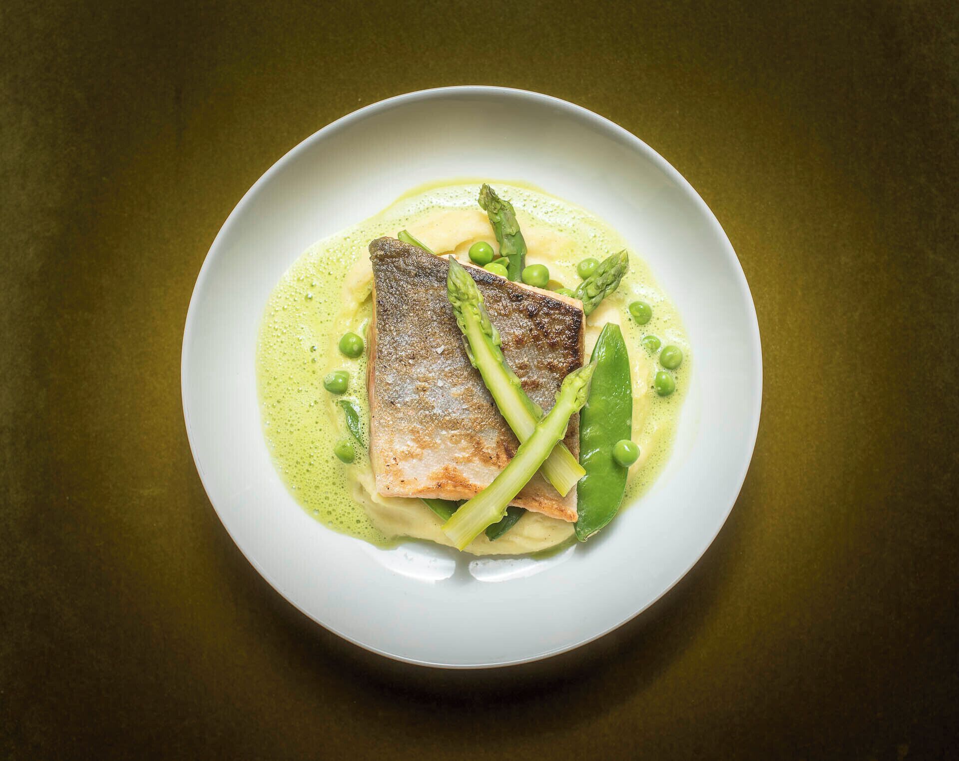 Ein weißer Teller mit Fisch, Spargel, Erbsen und einer grünen Soße auf dunklem Grund.