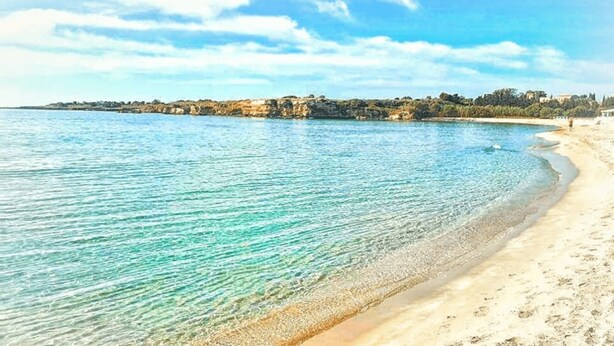 Una playa con aguas turquesas cristalinas, orilla arenosa y una costa rocosa distante con edificios.
