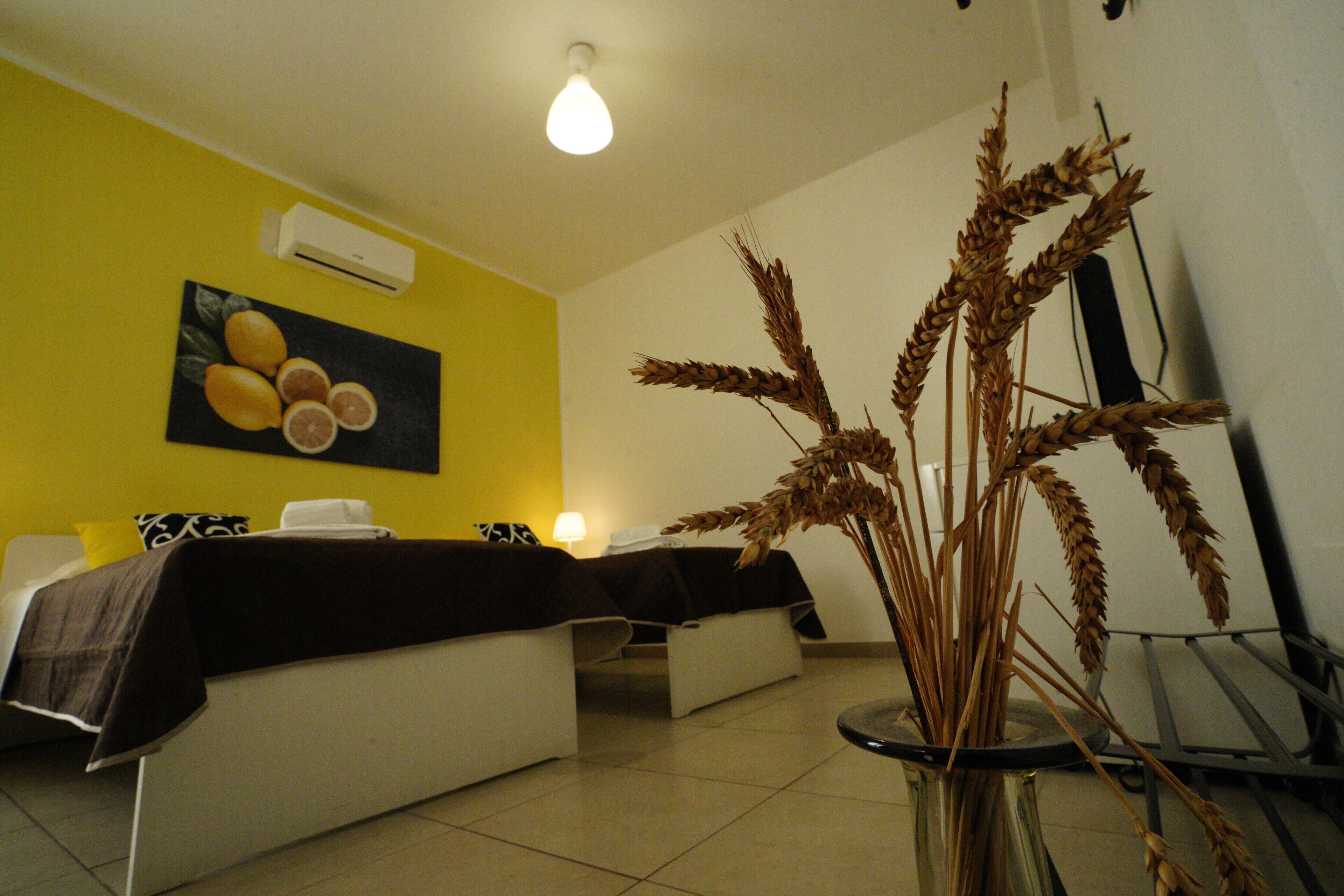 A room with a bed, a bedside table, a vase with dried plants, and a painting on the wall.