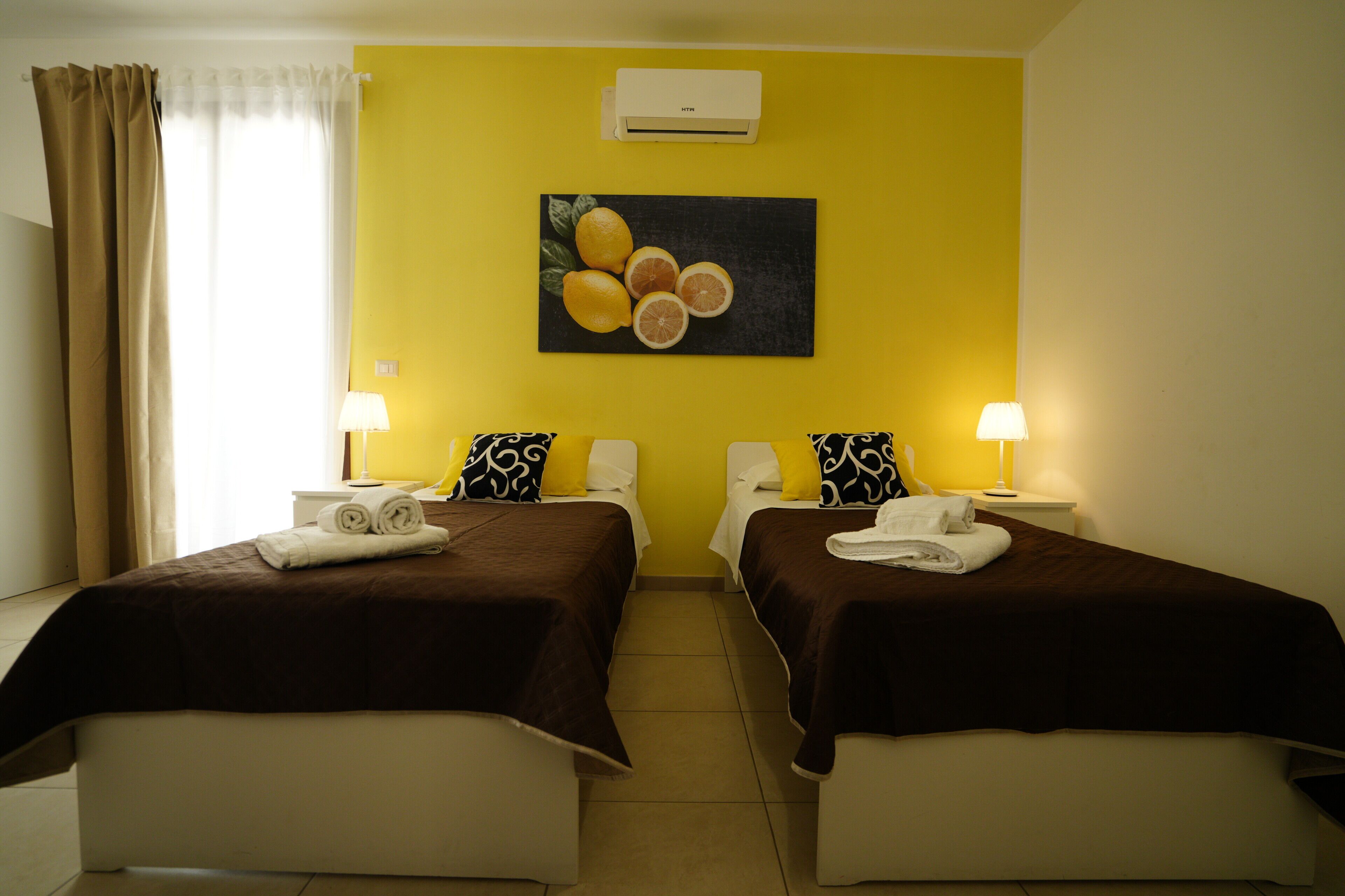 Une chambre d’hôtel avec deux lits, un mur jaune, un tableau représentant des agrumes et un appareil de climatisation.