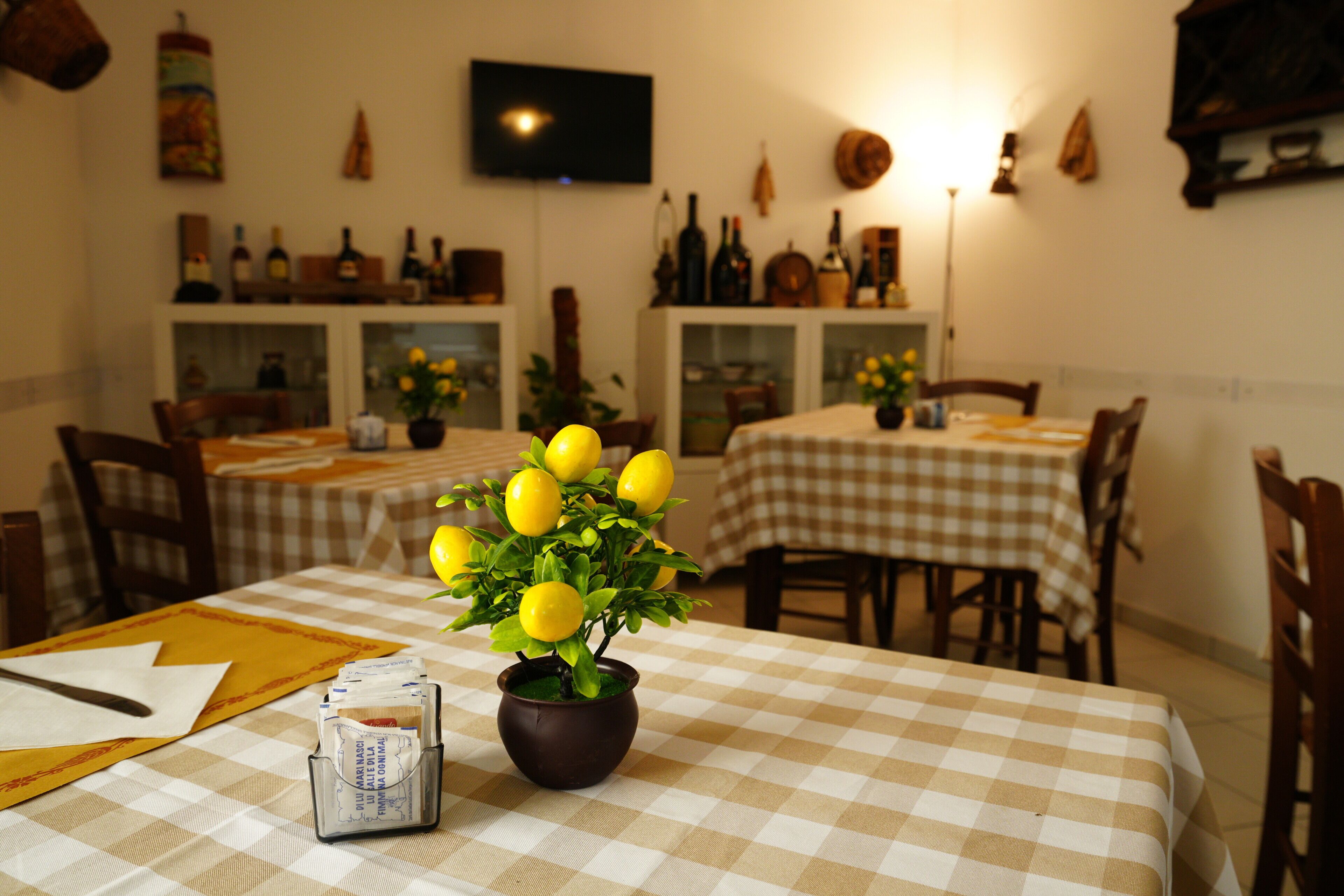 A dining area with checkered tablecloths, a potted lemon tree, and wine bottles on shelves.