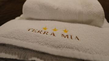 A neatly made bed with a white towel featuring 'TERRA MIA' and three stars, green and orange pillows, and a green wall with a blurred artwork.