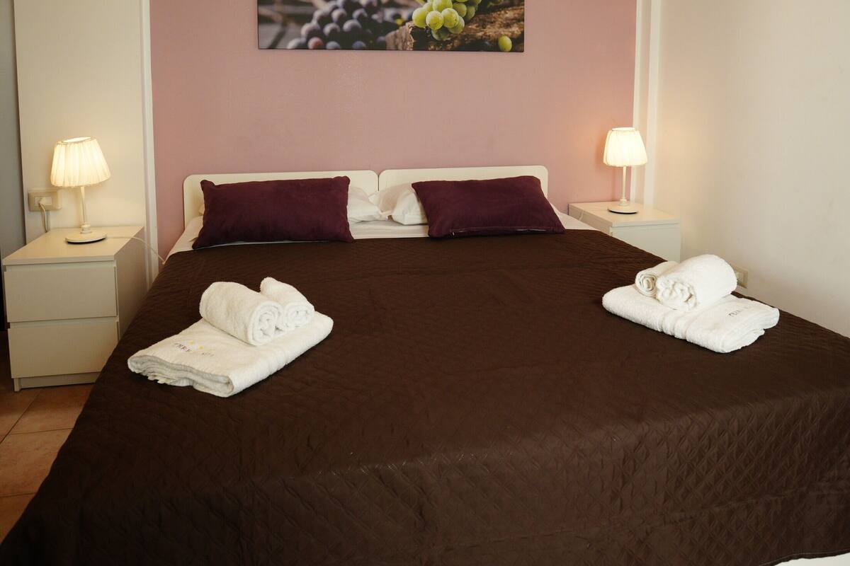 A neatly made bed with a brown bedspread, two white nightstands with lamps, a wall-mounted picture of grapes, and a pink wall.