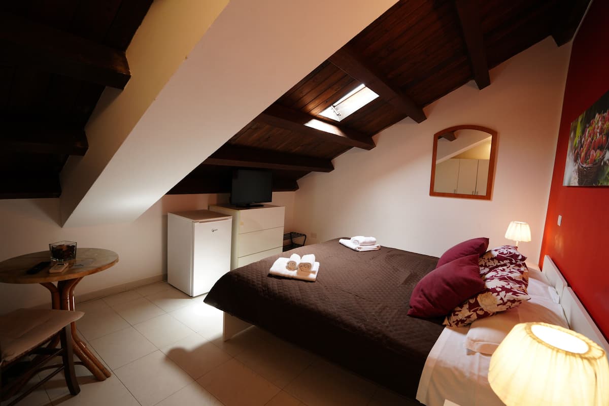 A bedroom with a sloped ceiling, a bed with a brown quilt, a bedside table with a lamp, a small desk with a chair, and a dresser.
