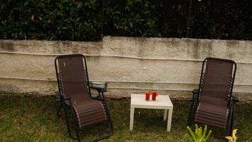 Two outdoor chairs with a small table between them, a white wall in the background, and a hedge.