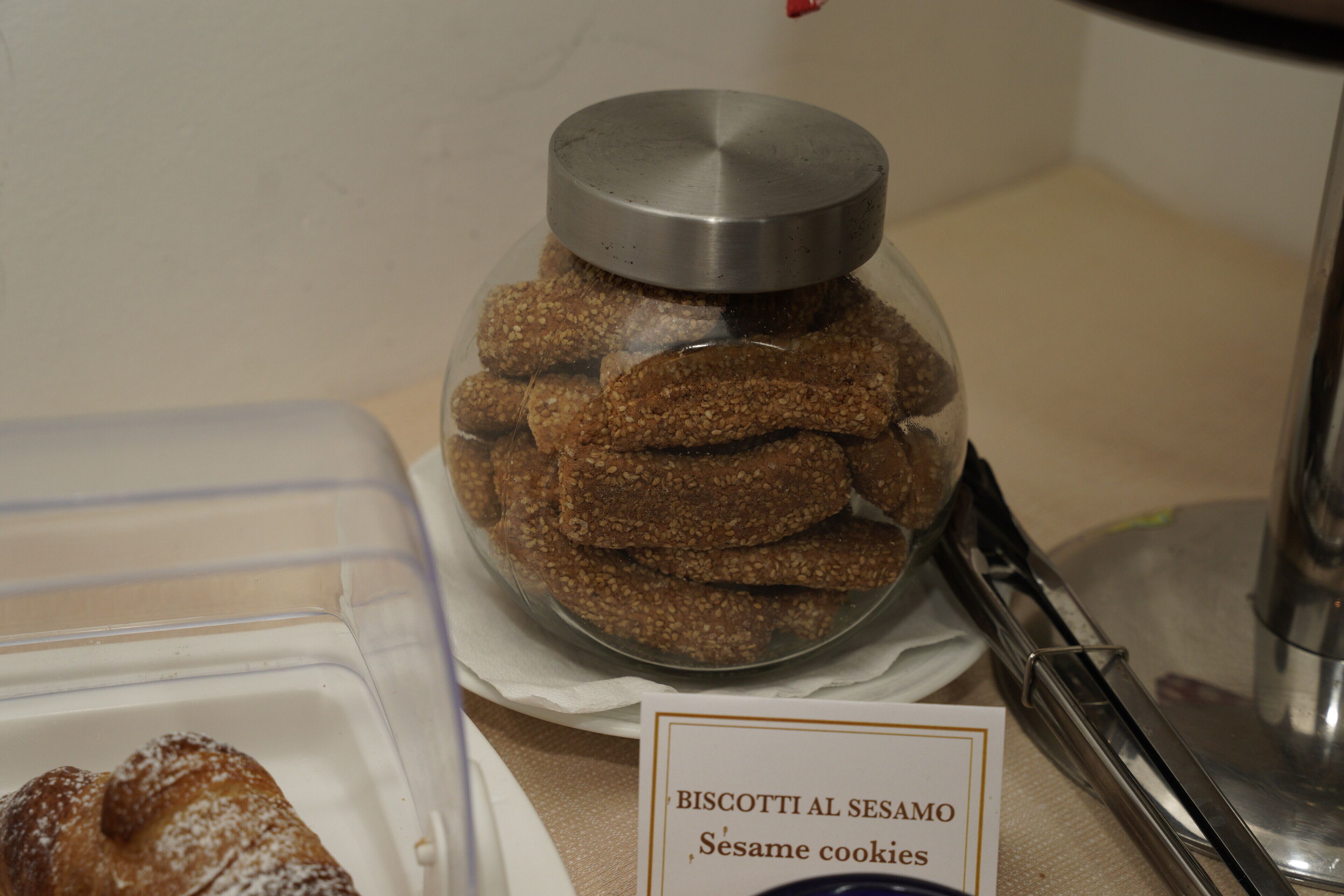 A container of sesame cookies labeled 'Biscotti al Sesamo' and a clear plastic container with a lid.
