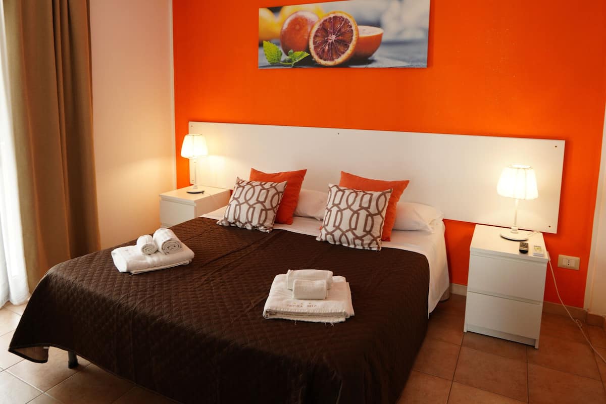 A hotel room with a bed, two bedside tables, a ceiling light, an air conditioning unit, and a fruit-themed painting on the wall.