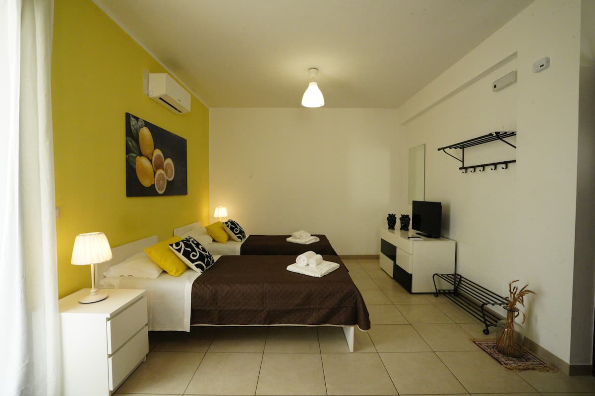 A hotel room with two beds, a TV, a small table, a lamp, a painting, and a wall-mounted shelf.
