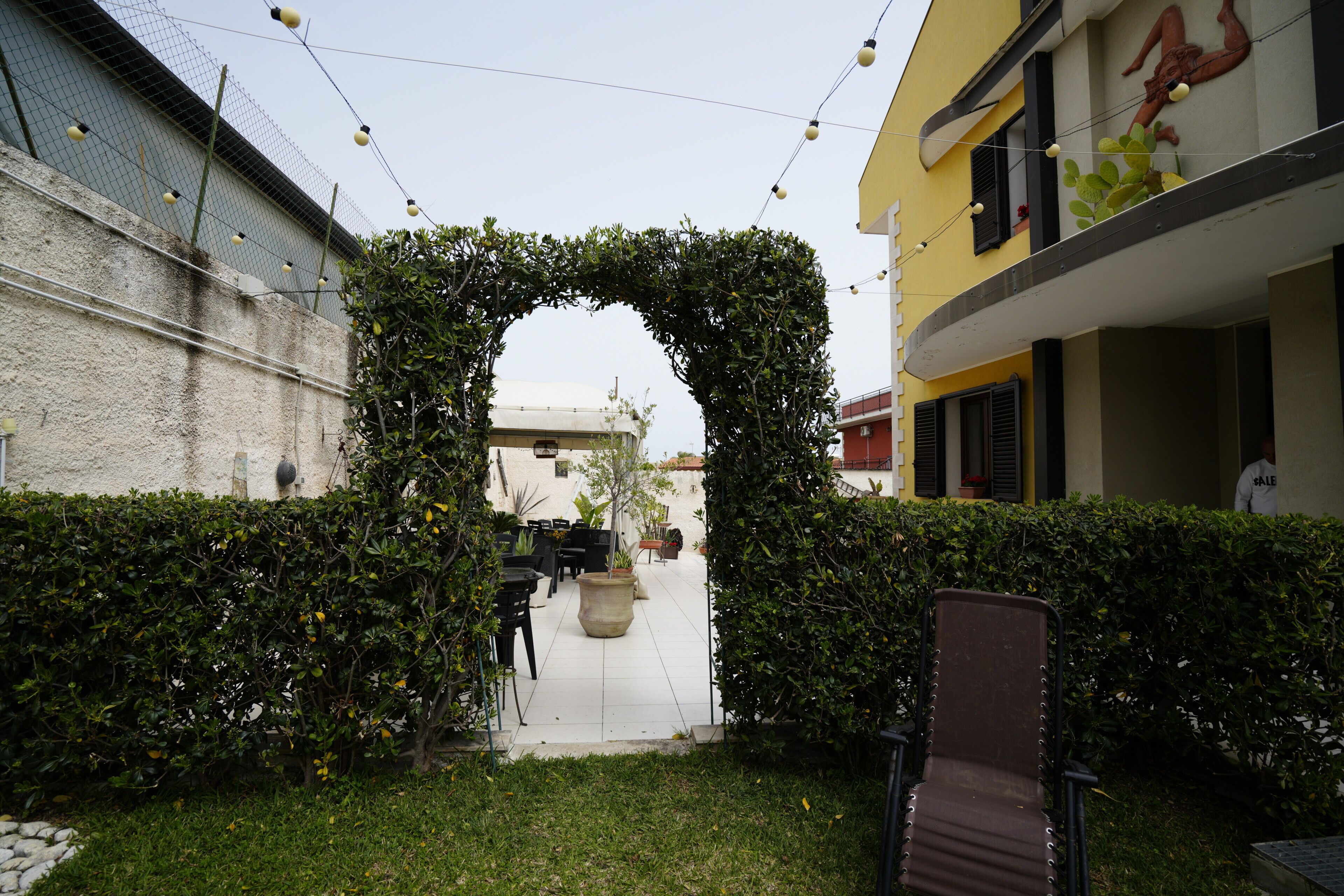 A courtyard with a covered seating area, a hedge archway, and a building with a balcony.