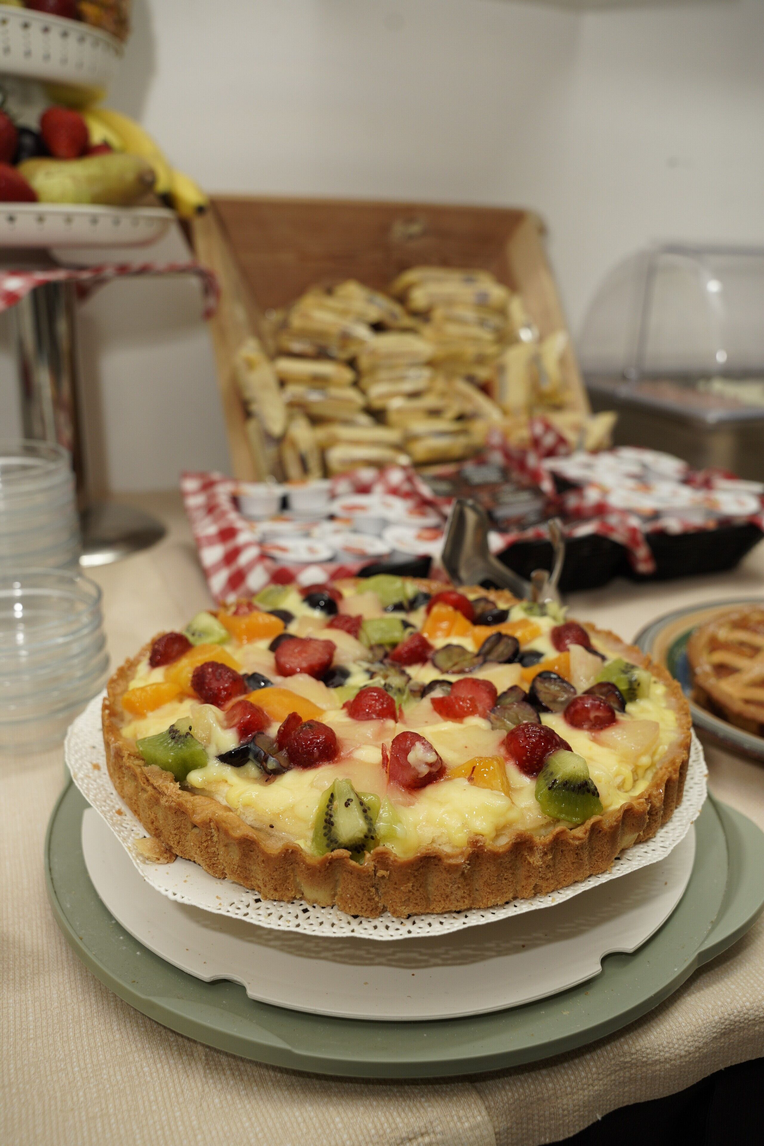 A fruit tart on a plate with a checkered cloth underneath, surrounded by other food items.