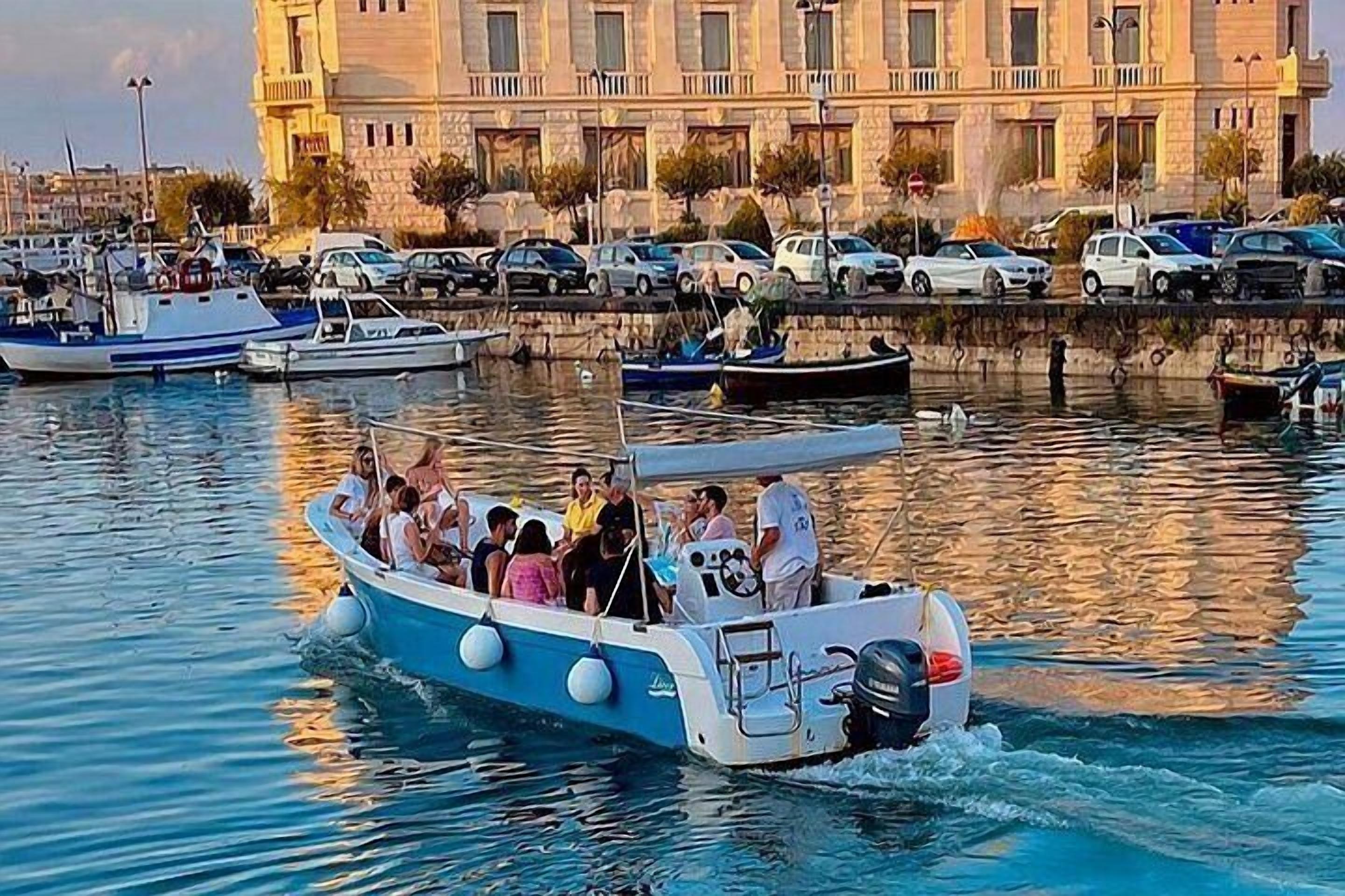 A boat with passengers, docked near a marina with cars and a building in the background.
