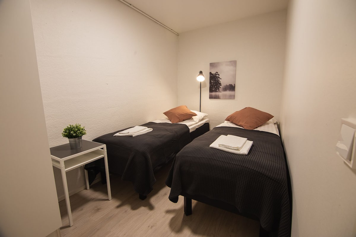 A small, well-lit room with two single beds, a bedside table with a plant, and a wall-mounted lamp.